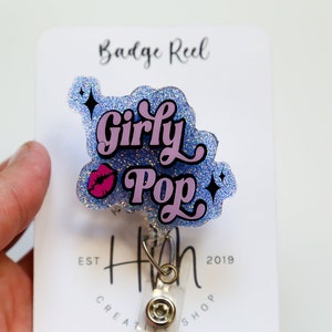 Op de afbeelding: Een glitter badge reel met de woorden "Girly Pop" in roze en zwart, met een roze lip afbeelding. De badge reel is bevestigd aan een witte kaart met de tekst "Badge Reel" en "H Creative Shop Est 2019".