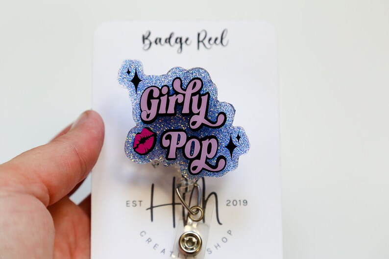 Op de afbeelding: Een glitter badge reel met de woorden "Girly Pop" in roze en zwart, met een roze lip afbeelding. De badge reel is bevestigd aan een witte kaart met de tekst "Badge Reel" en "H Creative Shop Est 2019".