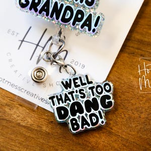 May include: A silver retractable badge holder with a black and white glitter charm that says "Well That's Too Dang Bad!" and a silver charm that says "Grandpa!"