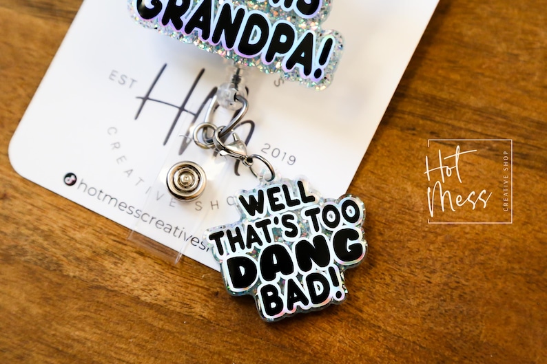 May include: A silver retractable badge holder with a black and white glitter charm that says "Well That's Too Dang Bad!" and a silver charm that says "Grandpa!"