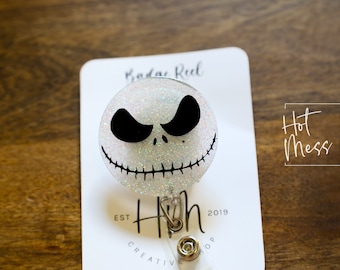May include: White glitter badge reel featuring a black and white Jack Skellington face design. The reel has a silver retractable cord and a silver clip. The text "Hot Mess Creative Shop" is visible in the background.