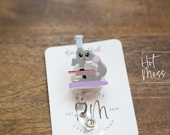 Può includere: Un microscopio di cartone animato grigio e rosa con una base viola, su una carta bianca con il testo "Hot Mess Creative Shop" e "Est. 2019".