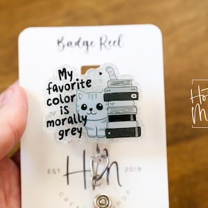 May include: A gray glitter badge reel with a cat and the text "My favorite color is morally grey". The cat is sitting on a stack of books. The badge reel is attached to a silver retractable badge holder.
