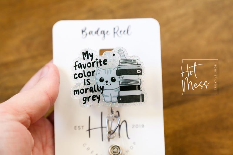 May include: A gray glitter badge reel with a cat and the text "My favorite color is morally grey". The cat is sitting on a stack of books. The badge reel is attached to a silver retractable badge holder.