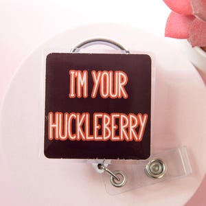 May include: A square, dark brown badge reel with the text "I'M YOUR HUCKLEBERRY" in orange lettering. The reel has a silver metal clip and a clear plastic strap. The background is a light pink.