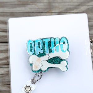 May include: A teal blue and white glitter badge reel with the word "ORTHO" and two broken bones with a bandage on one of the bones.