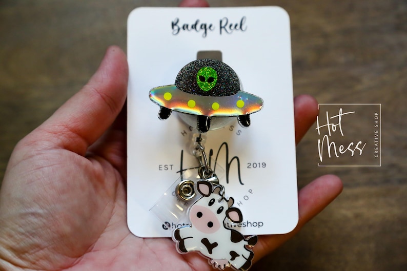 Puede incluir: Un carrete de insignia con un dise&ntilde;o de nave espacial alien&iacute;gena negro y verde brillante, con un dije de vaca. El carrete de insignia est&aacute; adjunto a una tarjeta blanca con el texto "Badge Reel" y "Hot Mess Creative Shop".