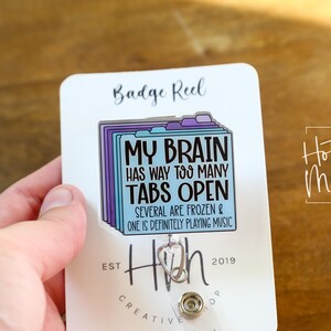 May include: A white badge reel with a rectangular design featuring a stack of folders in shades of blue and purple. The text reads "MY BRAIN HAS WAY TOO MANY TABS OPEN." The reel also has the text "SEVERAL ARE FROZEN & ONE IS DEFINITELY PLAYING MUSIC."