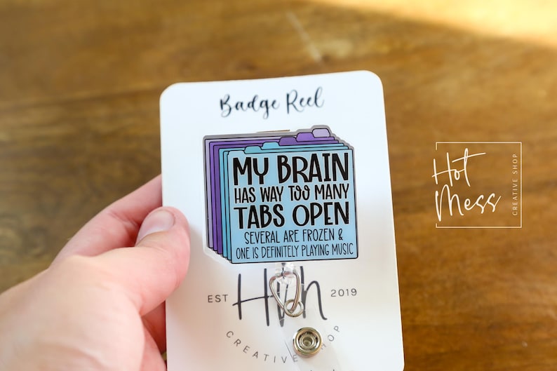May include: A white badge reel with a rectangular design featuring a stack of folders in shades of blue and purple. The text reads "MY BRAIN HAS WAY TOO MANY TABS OPEN." The reel also has the text "SEVERAL ARE FROZEN & ONE IS DEFINITELY PLAYING MUSIC."