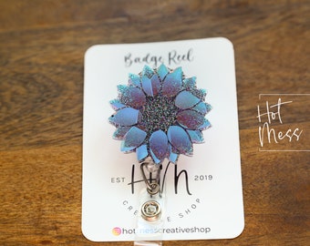 Puede incluir: Un carrete de insignia de girasol azul con purpurina con un cordón retráctil plateado y un clip plateado. El carrete está unido a una tarjeta blanca con el texto "Badge Reel", "Hot Mess Creative Shop", "Est 2019" y "Hotmesscreativeshop" impreso en ella.