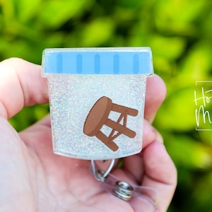 May include: A clear glitter badge holder with a blue stripe and a brown cartoon stool inside. The badge holder is attached to a retractable badge reel.