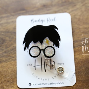 May include: A clear glitter badge reel with a black and white illustration of Harry Potter. The reel has a gold lightning bolt and the text "Hot Mess Creative Shop" is visible in the background.