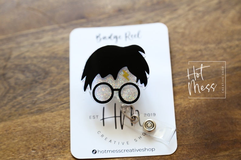 Puede incluir: Un carrete de insignia transparente con purpurina con una ilustraci&oacute;n en blanco y negro de Harry Potter. El carrete tiene un rayo dorado y el texto "Hot Mess Creative Shop" es visible en el fondo.
