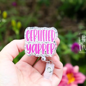 Puede incluir: Un portatarjetas con el texto "CERTIFIED YAPPER" en rosa brillante, delineado con purpurina plateada. El portatarjetas se sostiene en una mano sobre un fondo borroso de follaje verde y flores rosas.