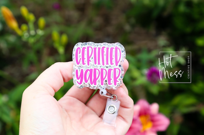 Puede incluir: Un portatarjetas con el texto "CERTIFIED YAPPER" en rosa brillante, delineado con purpurina plateada. El portatarjetas se sostiene en una mano sobre un fondo borroso de follaje verde y flores rosas.