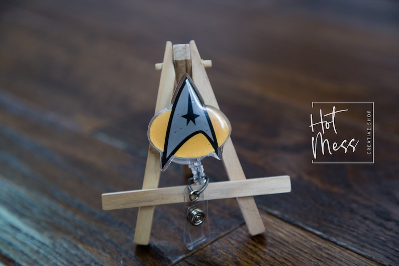 May include: A yellow and gray Star Trek insignia badge on a clear plastic retractable badge reel. The badge is displayed on a small wooden easel. The text "Hot Mess Creative Shop" is visible in the background.