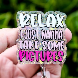 May include: A glittery badge with the words "RELAX I JUST WANNA TAKE SOME PICTURES" in white and pink. The badge is held in a hand against a blurred green background. The badge has a silver clip.