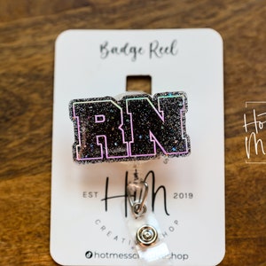 Op de afbeelding: Zwarte glitter badge reel met de letters "RN" in wit met een roze en groene omtrek. De badge reel is bevestigd aan een witte kaart met de tekst "Badge Reel" en "Hot Mess Creative Shop" erop gedrukt.