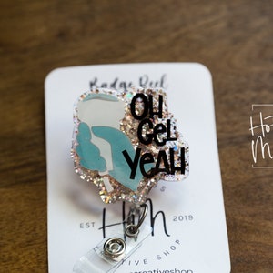 Könnte beinhalten: Ein glitzernder rosa-blauer Badge-Rollenhalter mit einer weißen und blauen Hand, die einen Haartrockner hält, und dem Text "Oh Gel Yeah". Der Badge-Rollenhalter ist an einer weißen Karte mit dem Text "Hot Mess Creative Shop" und "Est. 2019" befestigt.