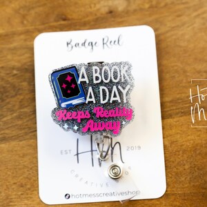 May include: A silver glitter badge reel with a pink and blue book charm that says "A Book A Day Keeps Reality Away". The charm is attached to a silver retractable badge holder. The badge reel is on a white card with the text "Badge Reel" and "Hot Mess Creative Shop".