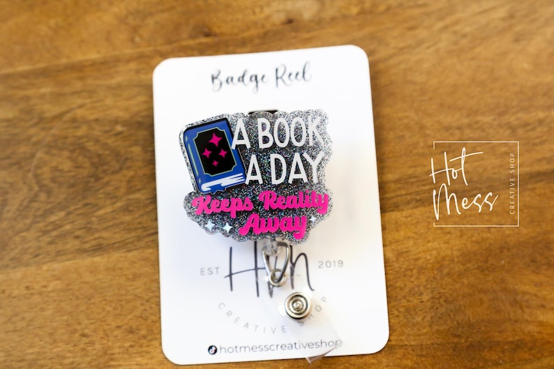 May include: A silver glitter badge reel with a pink and blue book charm that says "A Book A Day Keeps Reality Away". The charm is attached to a silver retractable badge holder. The badge reel is on a white card with the text "Badge Reel" and "Hot Mess Creative Shop".