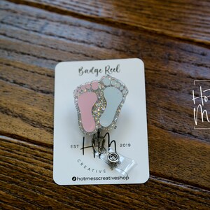May include: A silver badge reel with a pink and blue glitter baby footprint charm. The charm is attached to a clear plastic badge holder. The text "Badge Reel" is printed on a white card behind the charm. The text "Hot Mess Creative Shop" is printed on the card in a white square.