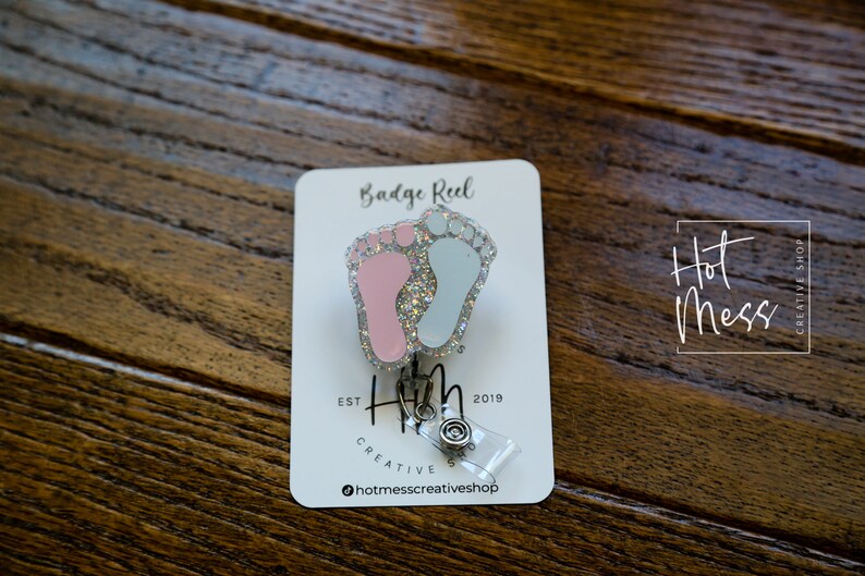 May include: A silver badge reel with a pink and blue glitter baby footprint charm. The charm is attached to a clear plastic badge holder. The text "Badge Reel" is printed on a white card behind the charm. The text "Hot Mess Creative Shop" is printed on the card in a white square.