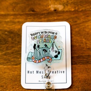 Op de afbeelding: Een doorzichtige badge reel met een cartoon opossum in een yogahouding op een blauwe mat. De tekst "Today's yoga pose is DOWNWARD SPIRAL" staat boven de opossum. De badge reel heeft een zilveren clip en een knop.