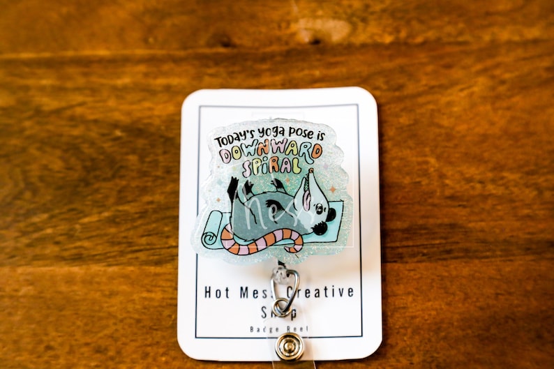 Op de afbeelding: Een doorzichtige badge reel met een cartoon opossum in een yogahouding op een blauwe mat. De tekst "Today's yoga pose is DOWNWARD SPIRAL" staat boven de opossum. De badge reel heeft een zilveren clip en een knop.