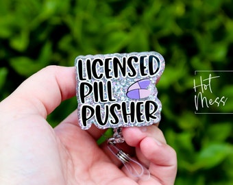 Może przedstawiać: Błyszczący identyfikator z napisem "LICENSED PILL PUSHER" w kolorze czarnym, obramowanym na biało. Dołączona jest grafika pigułki w kolorze fioletowym i różowym. Identyfikator jest trzymany w dłoni na rozmytym zielonym tle.