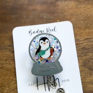 May include: A gray penguin snow globe badge reel with glitter inside. The penguin is holding a teal colored cup and a red book. The badge reel has a silver clip and a silver retractable cord. The text "Badge Reel" is printed on a white card behind the badge reel. The text "EST. HH 2019" is printed below the badge reel.
