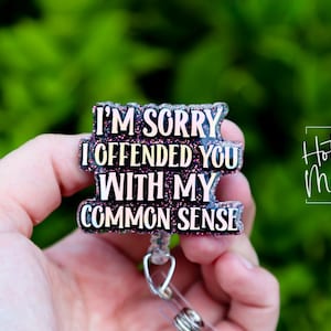 May include: A glittery badge reel with the text "I'M SORRY I OFFENDED YOU WITH MY COMMON SENSE" in white lettering. The badge reel is held in a hand against a blurred green background. The words "Hot Mess Creative Shop" are also visible.