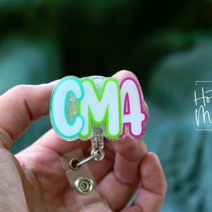 K&ouml;nnte beinhalten: Ein farbenfrohes Ausweis-Jojo mit den Buchstaben "CMA" in Wei&szlig;, umrandet von T&uuml;rkis, Gr&uuml;n und Pink. Das Ausweis-Jojo hat einen transparenten Kunststoffriemen und einen Metallclip. Der Text "Hot Mess Creative Shop" ist im Hintergrund zu sehen.