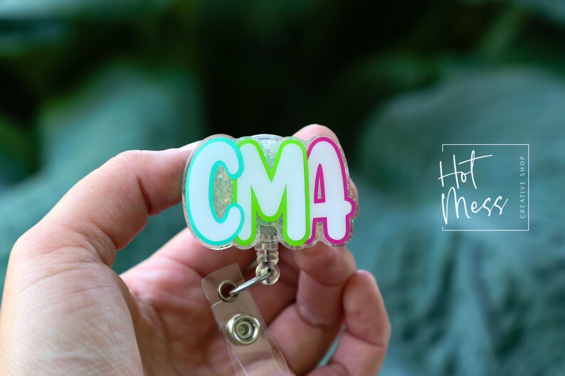 K&ouml;nnte beinhalten: Ein farbenfrohes Ausweis-Jojo mit den Buchstaben "CMA" in Wei&szlig;, umrandet von T&uuml;rkis, Gr&uuml;n und Pink. Das Ausweis-Jojo hat einen transparenten Kunststoffriemen und einen Metallclip. Der Text "Hot Mess Creative Shop" ist im Hintergrund zu sehen.