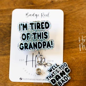 May include: A white card with the text "Badge Reel" and "I'm Tired of This Grandpa!" in black letters with glitter. A silver badge reel with a blue and white glitter charm that says "Well That's Too Dang Bad!" is attached to the card.