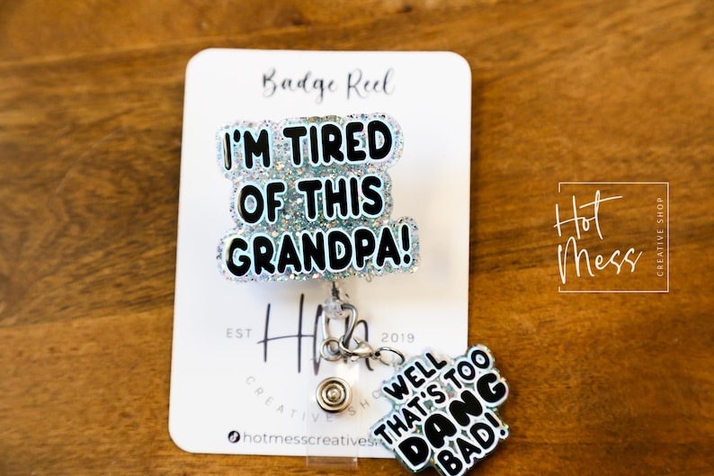 May include: A white card with the text "Badge Reel" and "I'm Tired of This Grandpa!" in black letters with glitter. A silver badge reel with a blue and white glitter charm that says "Well That's Too Dang Bad!" is attached to the card.