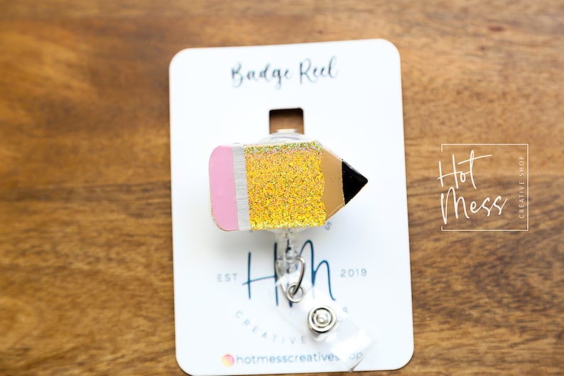 May include: A gold glitter pencil badge reel with a pink eraser and black tip. The reel is attached to a white card with the text "Badge Reel", "Hot Mess Creative Shop", "Est 2019", and "Hotmesscreatives.co".
