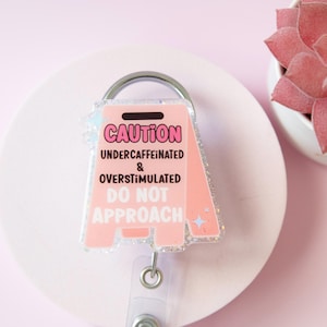 Puede incluir: Un portatarjetas rosa con purpurina en forma de señal de precaución. El texto dice CAUTION UNDERCAFFEINATED & OVERSTIMULATED DO NOT APPROACH en letras negras y blancas. El portatarjetas cuenta con un clip de metal plateado y una correa transparente.