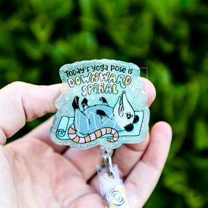Op de afbeelding: Een heldere, glinsterende badge reel met een cartoon opossum in een yogahouding op een blauwe mat. De tekst luidt "Today's yoga pose is DOWNWARD SPIRAL" in kleurrijke letters. De reel heeft een doorzichtige plastic clip.