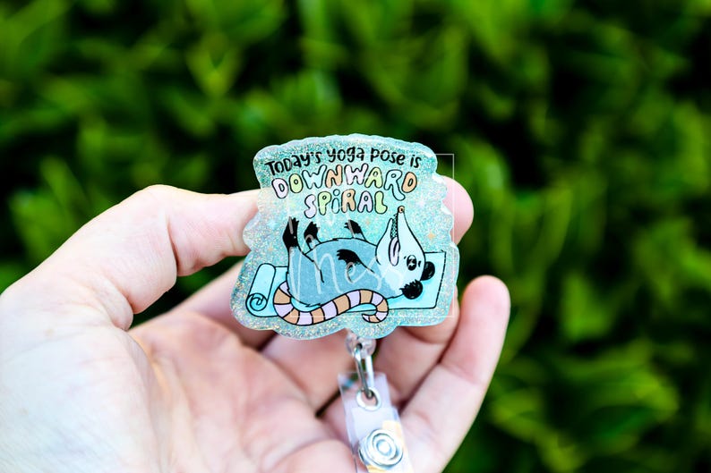Op de afbeelding: Een heldere, glinsterende badge reel met een cartoon opossum in een yogahouding op een blauwe mat. De tekst luidt "Today's yoga pose is DOWNWARD SPIRAL" in kleurrijke letters. De reel heeft een doorzichtige plastic clip.