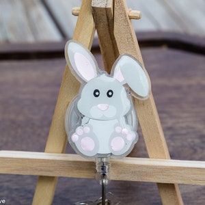 May include: A clear plastic retractable badge holder with a gray bunny design. The bunny has pink ears, a pink nose, and pink feet. The badge holder is attached to a silver chain.