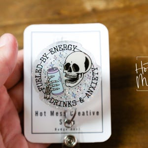 May include: A clear badge reel with a circular design featuring a skull, a skeleton hand holding a soda can, and the text "Fueled by Energy Drinks & Anxiety." The badge reel also includes the text "Hot Mess Creative Shop."