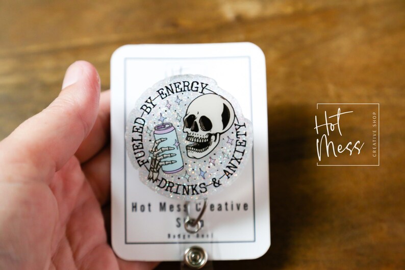 May include: A clear badge reel with a circular design featuring a skull, a skeleton hand holding a soda can, and the text "Fueled by Energy Drinks & Anxiety." The badge reel also includes the text "Hot Mess Creative Shop."