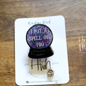 May include: A black and silver glitter badge reel with a black and silver glitter 8-ball charm. The charm has the text "I put a spell on you" on it. The badge reel is on a white card with the text "Hot Mess Creative Shop" on it.