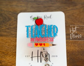 May include: A silver glitter badge holder with a blue and silver "Teacher in Progress" design, a red apple, a pencil, and two red hearts. The badge holder is attached to a clear retractable badge reel. The text "Hot Mess Creative Shop" is visible in the background.