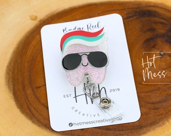 May include: A white glitter tooth with red, white, and blue hair wearing black sunglasses. The tooth is attached to a retractable badge holder with a silver clip. The text "Hot Mess Creative Shop" is printed on the card.