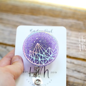 Puede incluir: Carrete de insignia de purpurina morada con un diseño geométrico holográfico y estrellas blancas. El texto "Hot Mess Creative Shop" es visible en el fondo.