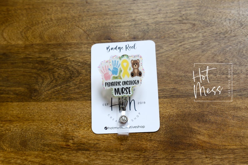 Peut inclure: Porte-badge blanc avec un fond paillet&eacute; et un insert en plastique transparent. L'insert pr&eacute;sente un ruban jaune, deux empreintes de mains et un ours en peluche brun. Le texte "Pediatric Oncology Nurse" est imprim&eacute; sur l'insert. Le texte "Hot Mess Creative Shop" est imprim&eacute; au bas du porte-badge.