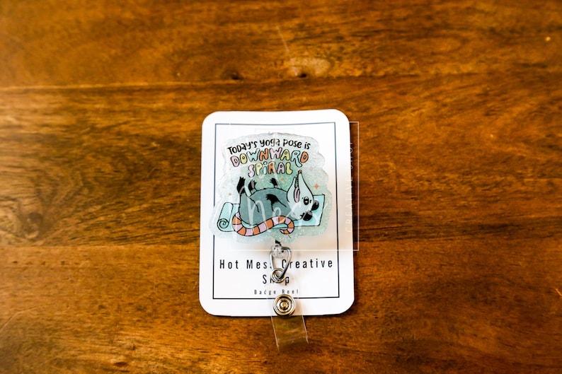 Op de afbeelding: Een witte badge reel met een cartoon illustratie van een rat in een yoga pose op een blauwe mat. De tekst "Today's yoga pose is downward spiral" staat boven de illustratie. De badge reel heeft de tekst "Hot Mess Creative Shop" en een doorzichtige plastic hoes.