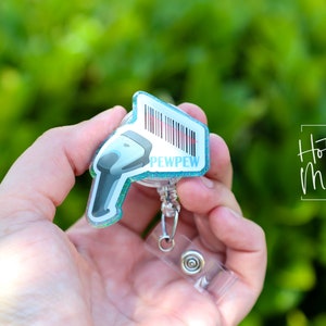 May include: A teal blue glitter badge holder with a white scanner design and the text "PEWPEW". The badge holder is attached to a clear plastic retractable badge reel.
